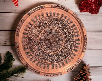 May include: A round copper tray with intricate black etched patterns. The tray features a central medallion design surrounded by detailed floral and geometric motifs. The tray's rim has a decorative border. The tray is displayed on a light wood surface.
