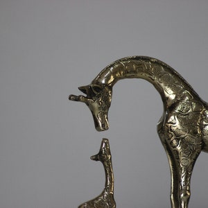 May include: A brass sculpture of a mother giraffe and her baby giraffe. The mother giraffe is standing on all fours and has her head bent down to look at her baby. The baby giraffe is standing on all fours and is looking up at its mother.