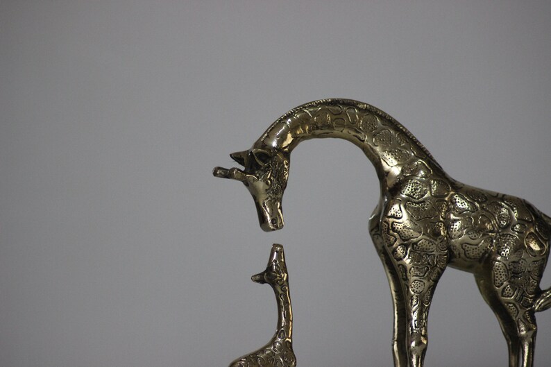 May include: A brass sculpture of a mother giraffe and her baby giraffe. The mother giraffe is standing on all fours and has her head bent down to look at her baby. The baby giraffe is standing on all fours and is looking up at its mother.