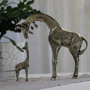 May include: Two brass giraffe figurines, one large and one small, standing on a light brown surface. The larger giraffe is facing left and the smaller giraffe is facing right. Both giraffes have a detailed, textured surface.