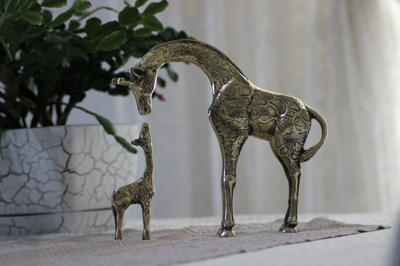 May include: Two brass giraffe figurines, one large and one small, standing on a light brown surface. The larger giraffe is facing left and the smaller giraffe is facing right. Both giraffes have a detailed, textured surface.