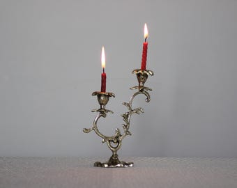 Puede incluir: Un candelabro plateado de dos velas con dos velas rojas encendidas. El candelabro tiene un diseño floral decorativo. Las velas arden con llamas amarillas brillantes. Está sobre una superficie gris.