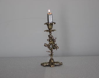 May include: A vintage silver candlestick with floral detailing and a lit candle. The candle is silver and has a small flame. The candlestick is ornate and stands on a four-leaf base.