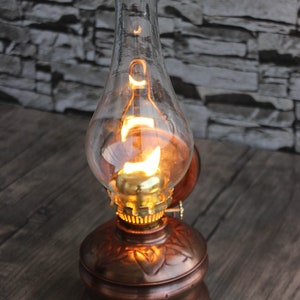 Puede incluir: Una l&aacute;mpara de aceite de cobre con una chimenea de vidrio transparente y una bombilla encendida en su interior. La l&aacute;mpara tiene un dise&ntilde;o decorativo en la base.