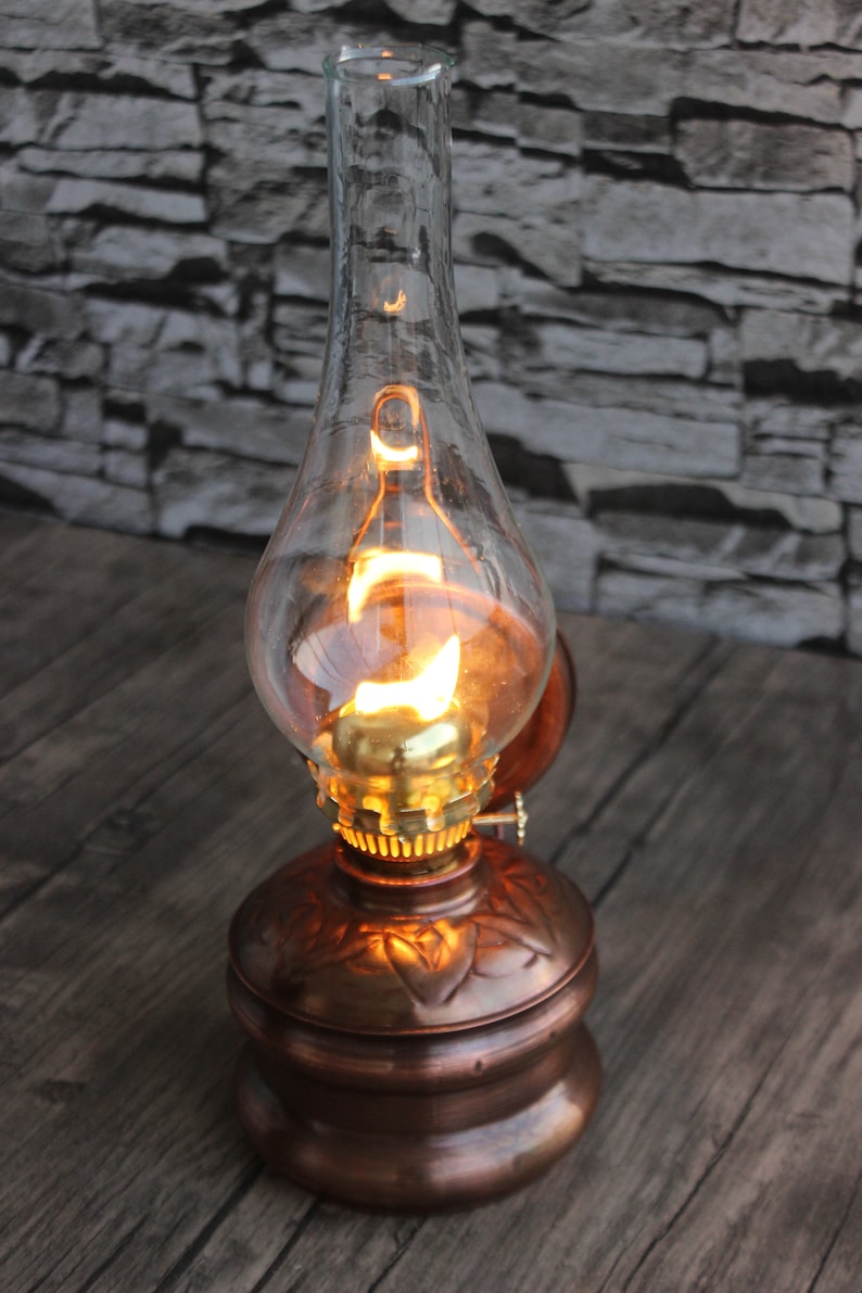 Puede incluir: Una l&aacute;mpara de aceite de cobre con una chimenea de vidrio transparente y una bombilla encendida en su interior. La l&aacute;mpara tiene un dise&ntilde;o decorativo en la base.