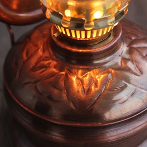 Puede incluir: Una l&aacute;mpara de aceite de cobre con un patr&oacute;n de hojas decorativas. La l&aacute;mpara tiene una parte superior de lat&oacute;n con una chimenea de vidrio y una mecha encendida.