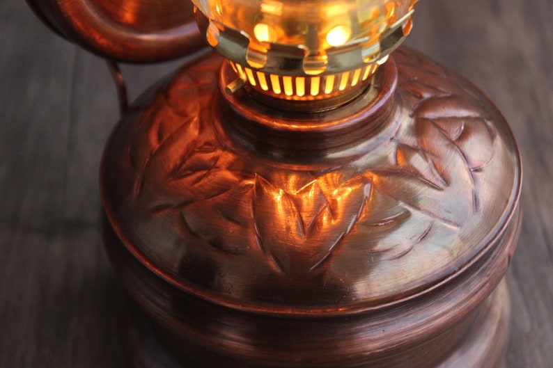 Puede incluir: Una l&aacute;mpara de aceite de cobre con un patr&oacute;n de hojas decorativas. La l&aacute;mpara tiene una parte superior de lat&oacute;n con una chimenea de vidrio y una mecha encendida.