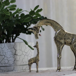 May include: Two brass giraffe figurines, one large and one small, standing on a table with a potted plant in the background. The larger giraffe has a detailed patterned design.