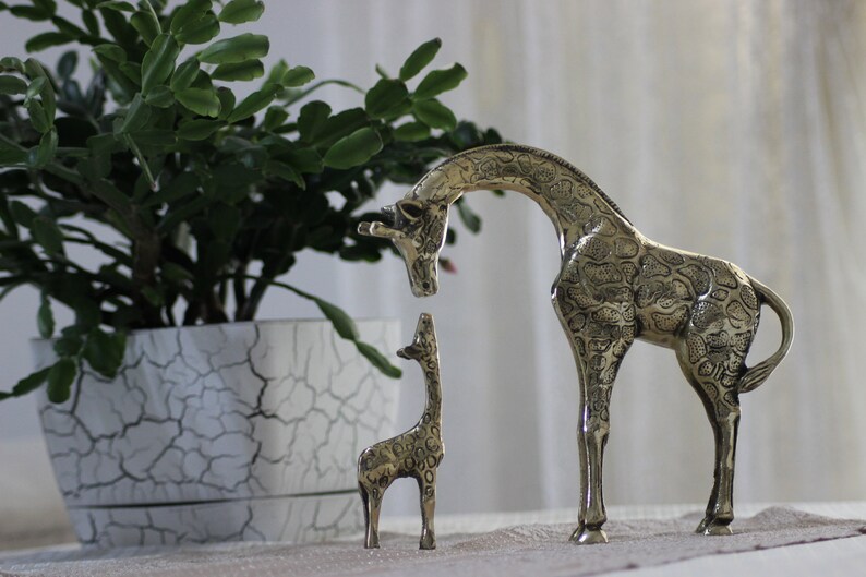 May include: Two brass giraffe figurines, one large and one small, standing on a table with a potted plant in the background. The larger giraffe has a detailed patterned design.