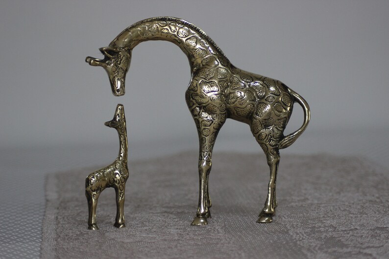 May include: A pair of brass giraffe figurines. The larger giraffe is standing with its head bent down, while the smaller giraffe is standing upright. Both giraffes have a detailed, textured surface.