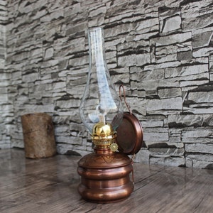 Puede incluir: Una l&aacute;mpara de aceite de cobre con una chimenea de vidrio y un soporte de pared. La l&aacute;mpara tiene un dise&ntilde;o decorativo y se encuentra en un entorno r&uacute;stico.