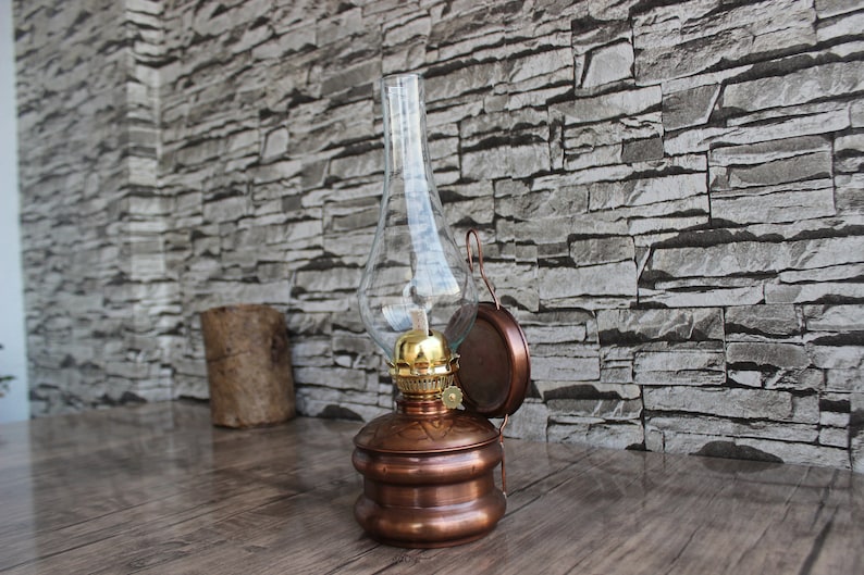 Puede incluir: Una l&aacute;mpara de aceite de cobre con una chimenea de vidrio y un soporte de pared. La l&aacute;mpara tiene un dise&ntilde;o decorativo y se encuentra en un entorno r&uacute;stico.