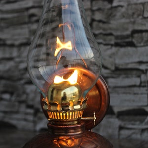 Puede incluir: Una l&aacute;mpara de aceite de estilo vintage con una pantalla de vidrio transparente y una base de cobre. La l&aacute;mpara est&aacute; encendida y proyecta un brillo c&aacute;lido.