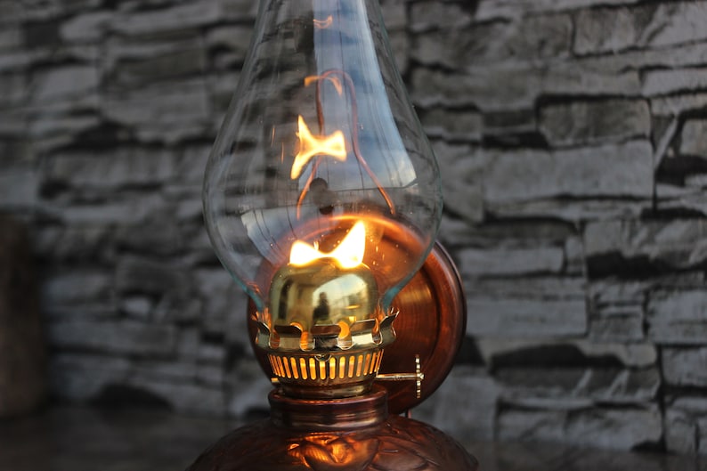 Puede incluir: Una l&aacute;mpara de aceite de estilo vintage con una pantalla de vidrio transparente y una base de cobre. La l&aacute;mpara est&aacute; encendida y proyecta un brillo c&aacute;lido.
