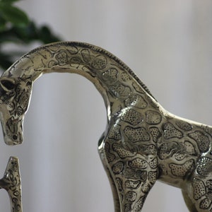 May include: A silver metal giraffe figurine with a detailed patterned design. The giraffe is standing on its hind legs with its head bent down.