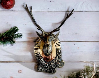 May include: A gold-toned metal deer head wall mount with intricate floral details around the edges. The deer head is mounted on a white wooden wall.