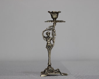 May include: A silver candlestick with a decorative figure of a woman holding a flower. The candlestick has a round base with three legs.