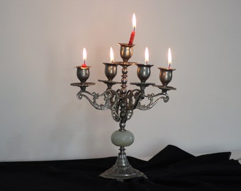 May include: An ornate silver candelabra with six lit candles. The candelabra has a decorative base and arms, with a central column and a light-colored stone. The candles are red and have visible flames.