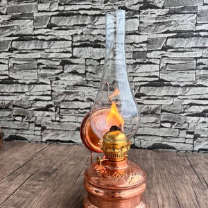 Puede incluir: Una l&aacute;mpara de aceite de cobre con una chimenea de vidrio y una llama en su interior. La l&aacute;mpara tiene un dise&ntilde;o decorativo en la base y un asa en el lateral.