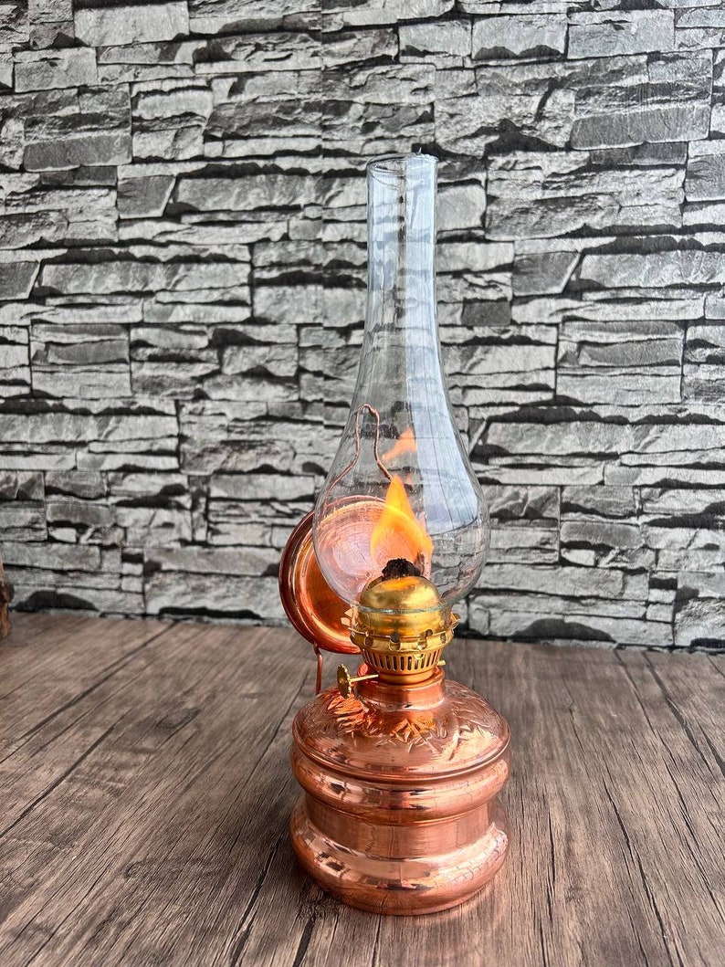 Puede incluir: Una l&aacute;mpara de aceite de cobre con una chimenea de vidrio y una llama en su interior. La l&aacute;mpara tiene un dise&ntilde;o decorativo en la base y un asa en el lateral.