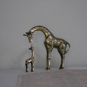 May include: Two gold-colored metal giraffe figurines. The larger giraffe is standing with its head bent down, looking at the smaller giraffe. The smaller giraffe is standing upright.