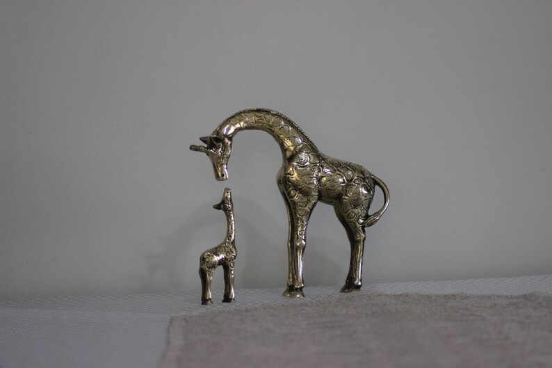 May include: Two gold-colored metal giraffe figurines. The larger giraffe is standing with its head bent down, looking at the smaller giraffe. The smaller giraffe is standing upright.