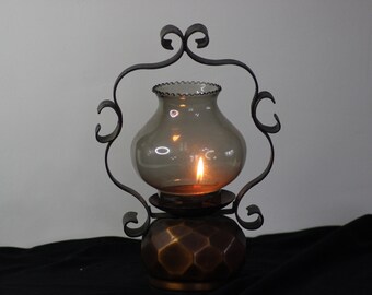 May include: A vintage candle lantern with a lit flame inside a smoky glass globe. The lantern is supported by a decorative wrought iron frame and a copper-toned base with a geometric pattern. The overall design is ornate.