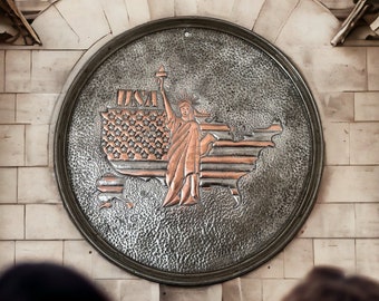 May include: A circular metal plaque with a raised design of the Statue of Liberty standing on a map of the United States. The plaque is silver with a copper-colored design and the word "USA" is embossed at the top.