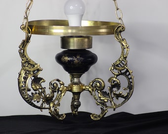 May include: Ornate brass chandelier with a black and gold center piece. The chandelier features intricate floral and scrollwork designs, and a white lightbulb. The fixture hangs from a chain.