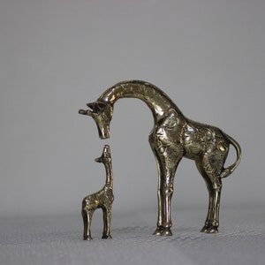 May include: Two brass giraffe figurines, one larger than the other, standing on a white surface. The larger giraffe is looking down at the smaller giraffe.