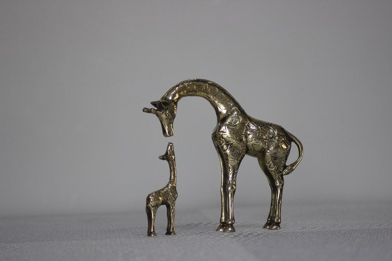May include: Two brass giraffe figurines, one larger than the other, standing on a white surface. The larger giraffe is looking down at the smaller giraffe.