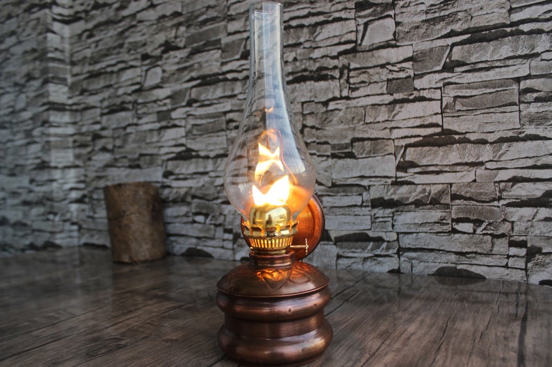 Puede incluir: Una l&aacute;mpara de aceite de pared de cobre con una pantalla de vidrio transparente y una bombilla encendida en su interior. La l&aacute;mpara est&aacute; montada en una pared de ladrillo.