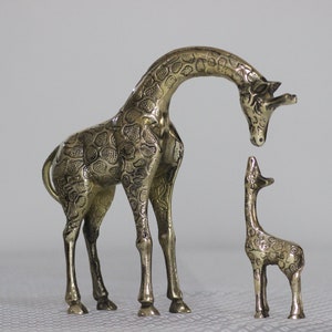 May include: Two gold-colored metal giraffe figurines. The larger giraffe is standing with its head bent down, looking at the smaller giraffe. The smaller giraffe is standing upright with its head tilted back, looking up at the larger giraffe.