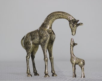 May include: Two gold-colored metal giraffe figurines. The larger giraffe is standing with its head bent down, looking at the smaller giraffe. The smaller giraffe is standing upright with its head tilted back, looking up at the larger giraffe.