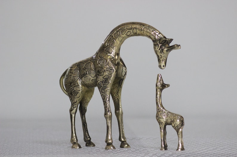 May include: Two gold-colored metal giraffe figurines. The larger giraffe is standing with its head bent down, looking at the smaller giraffe. The smaller giraffe is standing upright with its head tilted back, looking up at the larger giraffe.