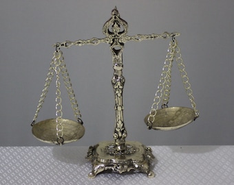 May include: A silver decorative scale with ornate details and two hanging bowls. The scale is on a white surface.