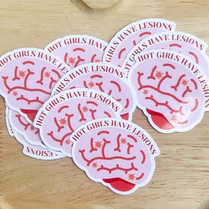 May include: A collection of stickers featuring a pink brain design with the text "HOT GIRLS HAVE LESIONS" in a playful font. The stickers are white-bordered and arranged on a wooden tray.