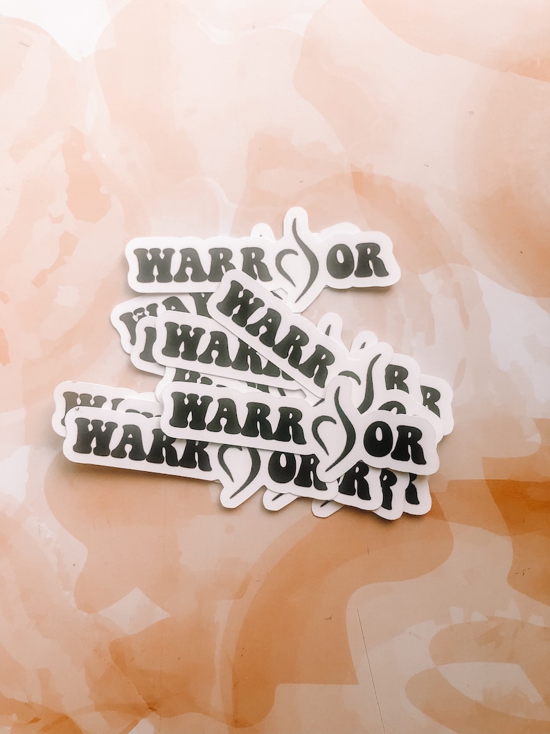 May include: A stack of white stickers with black text that reads "Warrior" with a stylised heart symbol. The stickers are on a light brown background.
