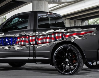 Könnte beinhalten: Ein grauer Pickup mit einem zerrissenen Aufkleber mit der amerikanischen Flagge an der Seite. Der Wagen hat schwarze Räder und eine schwarze Stoßstange.