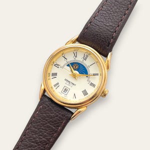 Puede incluir: Un reloj de pulsera dorado con esfera color crema, números romanos y correa de cuero texturizado marrón oscuro. La esfera incluye un indicador de fase lunar con gráfico de sol y luna, una ventana de fecha y el texto CONSTANT QUARTZ.