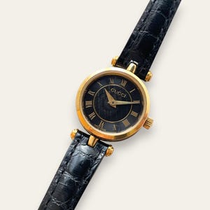 May include: A Gucci wristwatch featuring a round gold-tone case and a black dial with gold Roman numeral hour markers. The watch has a black leather strap with a textured, embossed finish and gold-tone hardware connecting the strap to the case.