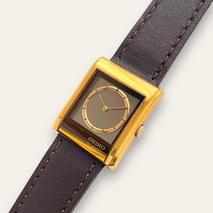 Puede incluir: Un reloj Seiko Quartz dorado con una esfera cuadrada y una correa de cuero marrón. La esfera del reloj es de color gris oscuro con manecillas doradas y un bisel dorado. La correa es de color marrón oscuro con costuras visibles.