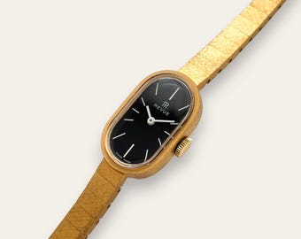 May include: A vintage gold wristwatch with a black oval face. The watch has a gold strap and a small gold crown. The watch face has white hands and markers, with the word "REVUE" printed on the face. The watch is marked "SWISS MADE".
