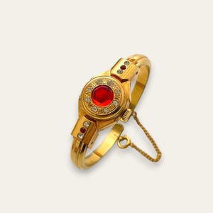 May include: A gold-toned bracelet with a large, round, red gemstone at its center, surrounded by smaller clear stones. The bracelet features additional red and clear stones and a safety chain. The design is ornate and vintage-inspired.