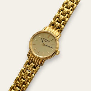 May include: A gold-tone analogue wristwatch featuring a round champagne-coloured dial with thin gold hour markers and the Longines logo. The watch has a polished gold-tone metal link bracelet with a textured, multi-link design and a side crown.