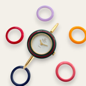 Puede incluir: Un reloj Gucci de cuarzo con esfera blanca, agujas doradas y bisel a rayas verdes y rojas. El reloj está rodeado de cinco anillos intercambiables de colores: rojo, morado, naranja, rosa y azul.