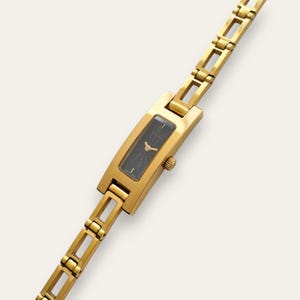 Puede incluir: Un reloj de pulsera dorado con una esfera negra rectangular y una correa de eslabones dorados. La esfera muestra agujas doradas y la marca "GUCCI". Este elegante reloj es un diseño clásico, adecuado para el uso diario.