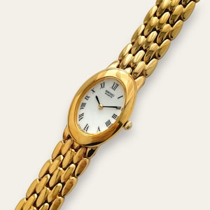 Può includere: Un orologio da polso Seiko Quartz color oro con cassa ovale e quadrante bianco con numeri romani neri. L'orologio presenta un cinturino a maglie in metallo color oro lucido con segmenti arrotondati e tre lancette sottili.