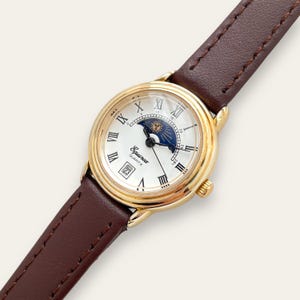 Peut inclure: Une montre Equinox Quartz dorée avec un cadran blanc, des chiffres romains et un affichage de la date. Le bracelet est en cuir marron. Le cadran de la montre présente un motif de soleil et de lune. Un accessoire classique.