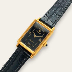 May include: A gold-toned rectangular Nivada watch with a black face and gold hands. The watch features Roman numerals and the brand name. The watch has a black leather band.